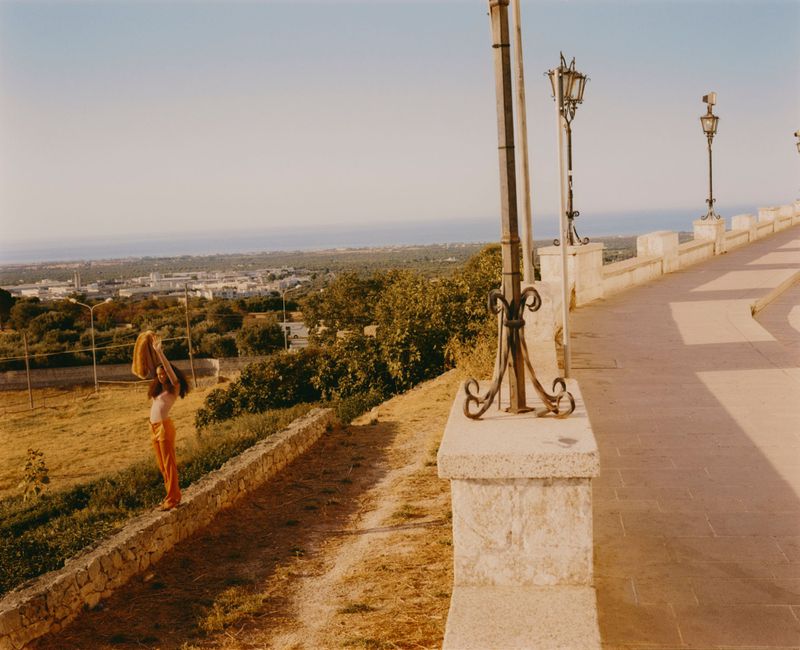 Woman in retro orange pants stands by stone wall with town and sea views, holding yellow shawl in outdoor, sunny, summertime setting.