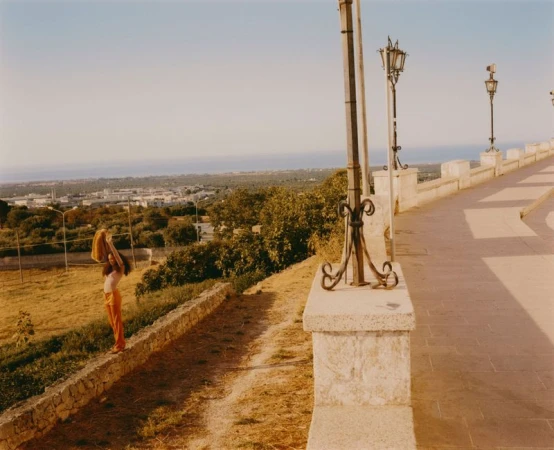 Woman in retro orange pants stands by stone wall with town and sea views, holding yellow shawl in outdoor, sunny, summertime setting.