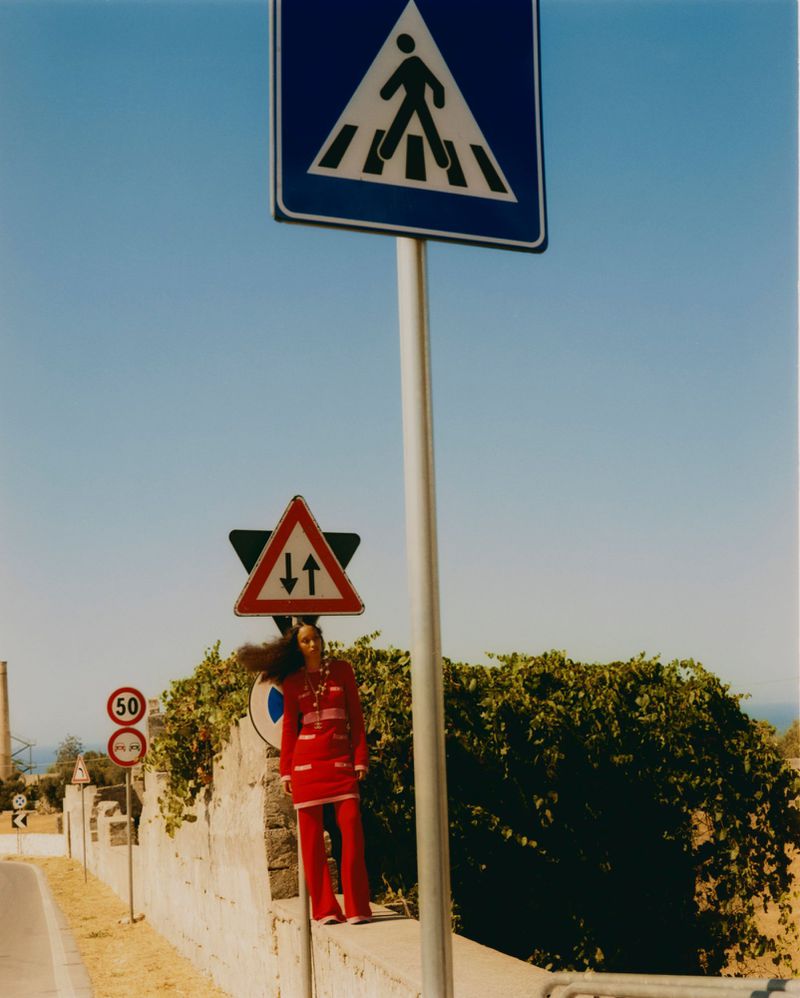 A woman in a red suit stands beside road signs and a stone wall against a bright blue sky in an outdoor daytime fashion shot.
