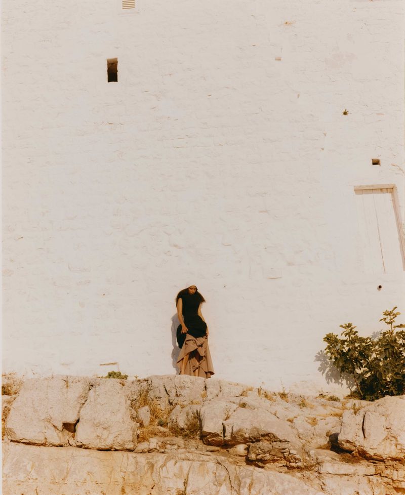 Woman in long skirt stands on rocky cliff in front of white stone wall under bright sunlight, with small green bush to the right.