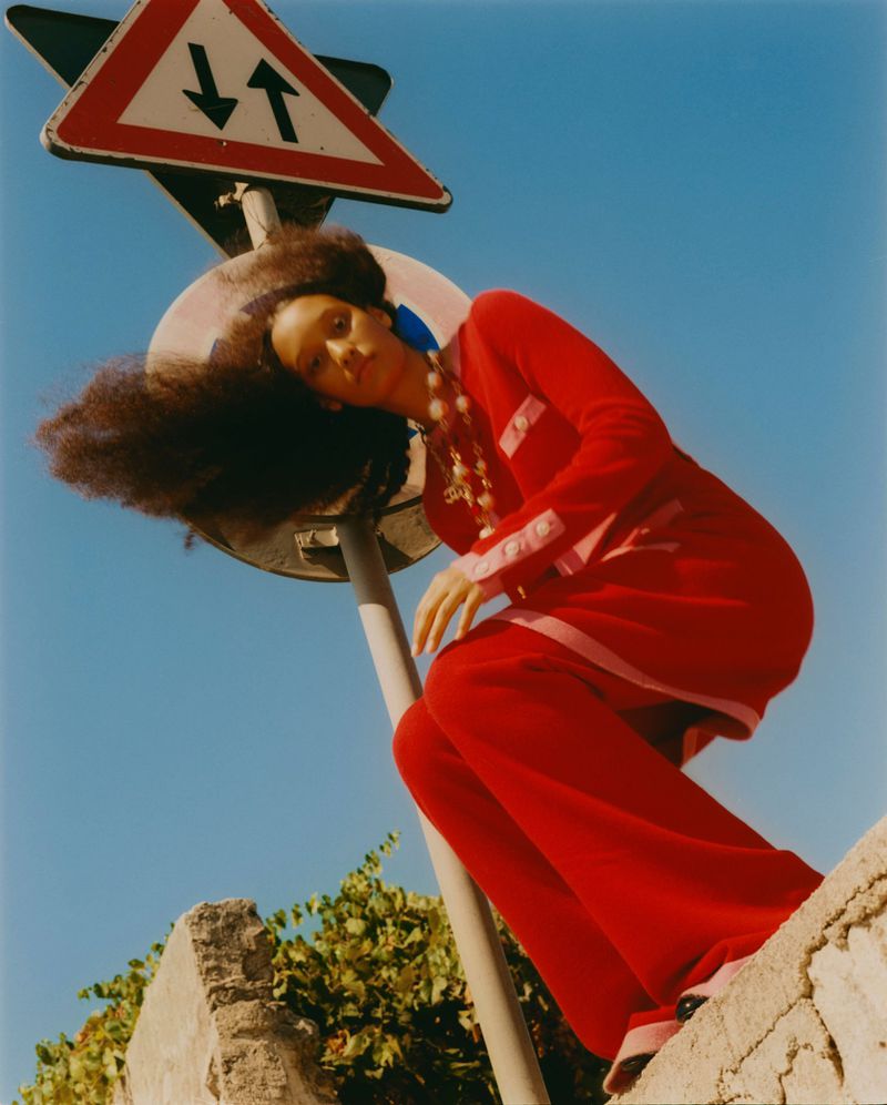 A woman in a red outfit poses by a road sign. Her hair is voluminous, and she wears large earrings. She poses outdoors against a blue sky.