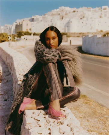 Model seated on a stone wall in front of a white hilltop town wearing a textured grey coat and pink shoes.