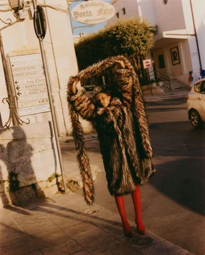 Model in a fluffy faux fur coat and bright red boots posing on a sunlit historic town street for L'Officiel Singapore.
