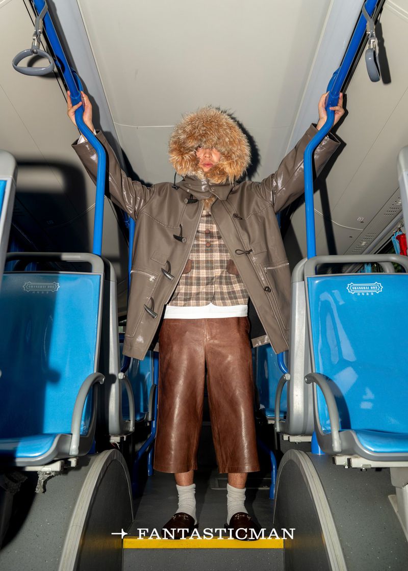 Model wearing a fur hat and leather jacket on a bus. The person is standing and holding onto the blue rails, looking stylish.