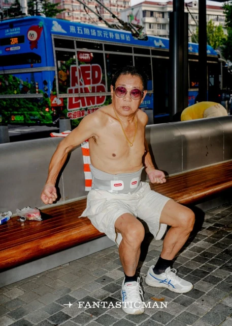 Bare-chested older man with sunglasses and compression belt sitting on a wooden bench in an urban environment, with a bus in background.