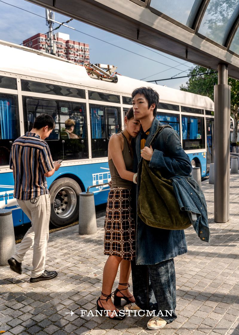 Young couple embrace at bus stop in urban setting. Man in denim coat and woman in patterned skirt, with bus and city buildings visible.