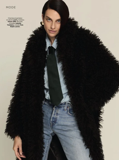 A woman in a black shaggy coat, light blue shirt, green tie, and light blue jeans poses in a studio. Fashion editorial portrait.