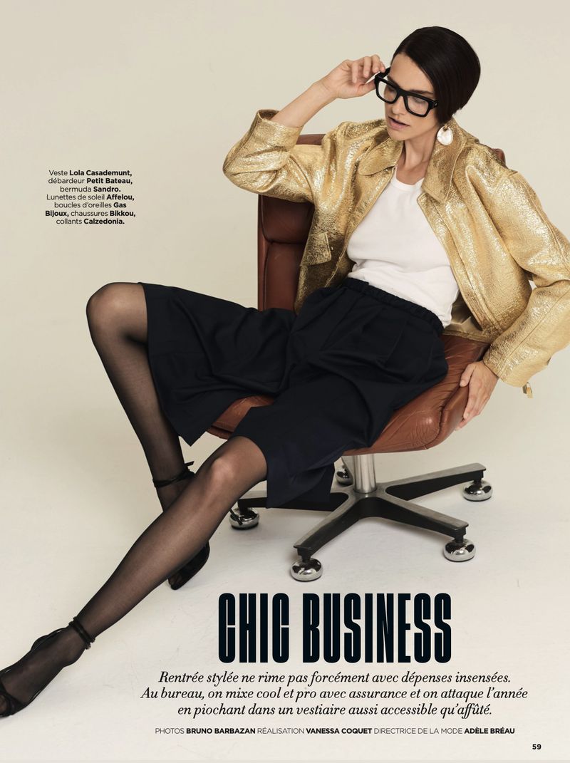 A woman in a gold jacket and black shorts sits on an office chair, wearing black glasses in an indoor setting, with a fashion editorial feel.