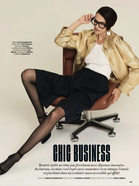A woman in a gold jacket and black shorts sits on an office chair, wearing black glasses in an indoor setting, with a fashion editorial feel.