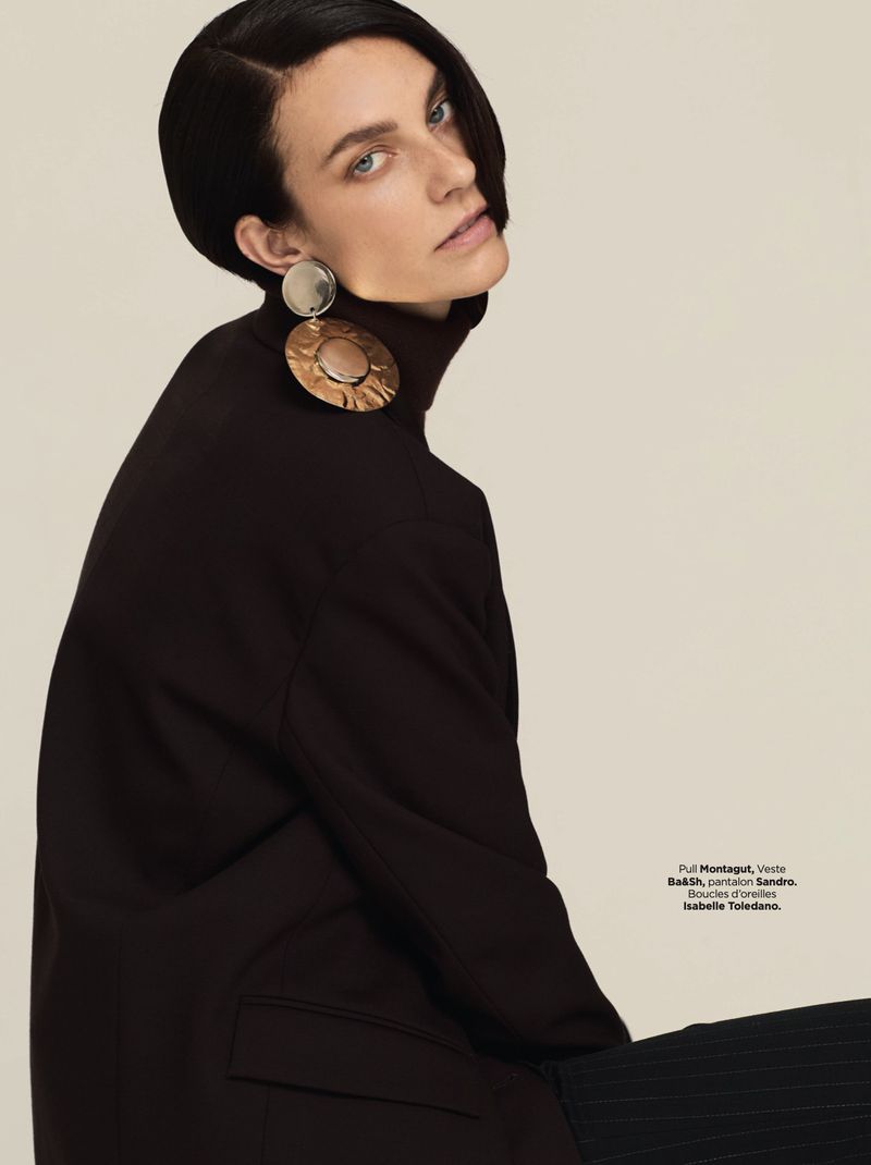 A fashion shot of an adult female model with short, dark hair, wearing large earrings and a dark turtleneck against a neutral background.