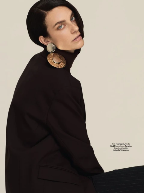 A fashion shot of an adult female model with short, dark hair, wearing large earrings and a dark turtleneck against a neutral background.