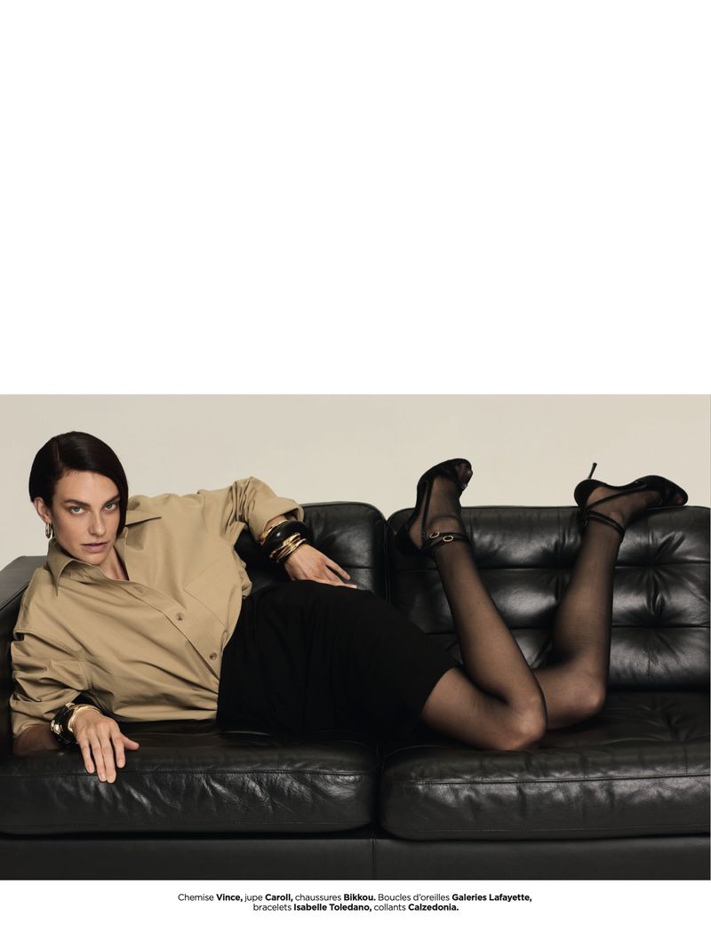Fashion shot of a woman reclining on a black couch wearing a skirt, tan blouse, black hosiery, and heels in an indoor environment.