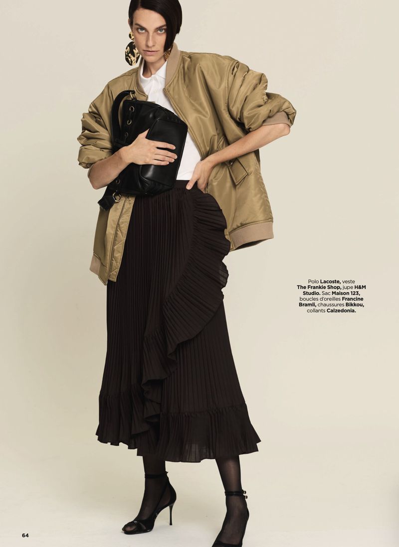 Fashion editorial shot of a woman in a bomber jacket and pleated skirt, holding a black bag, accessorized with large earrings.