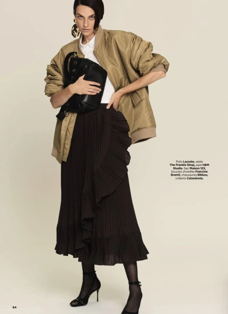 Fashion editorial shot of a woman in a bomber jacket and pleated skirt, holding a black bag, accessorized with large earrings.