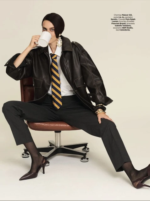Stylish woman in leather jacket and heels drinks coffee, seated on an office chair. Fashion-forward pose in studio setting.
