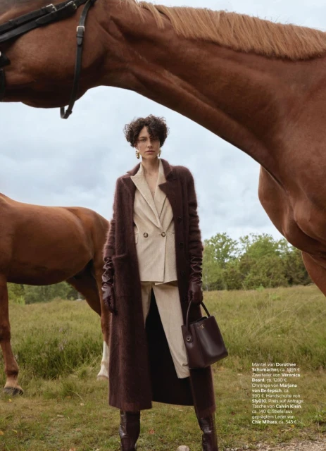 Woman in an overcoat and suit stands outdoors with a brown leather purse. Two brown horses stand in the background on a grassy field.