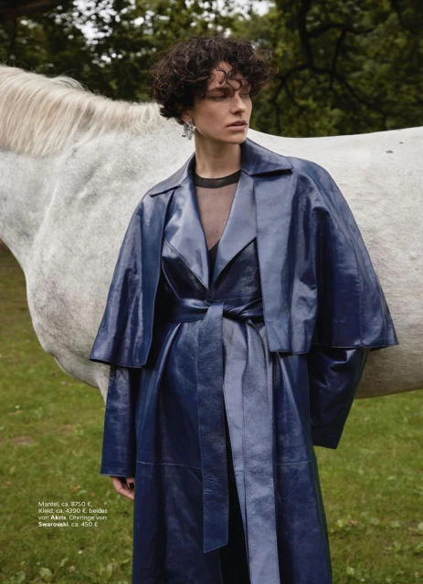 A woman in a blue belted coat poses with a white horse in a green field, creating a striking fashion shot.
