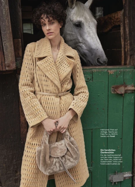 A woman wearing a patterned coat poses next to a white horse peering from a weathered green barn door in an outdoor setting.