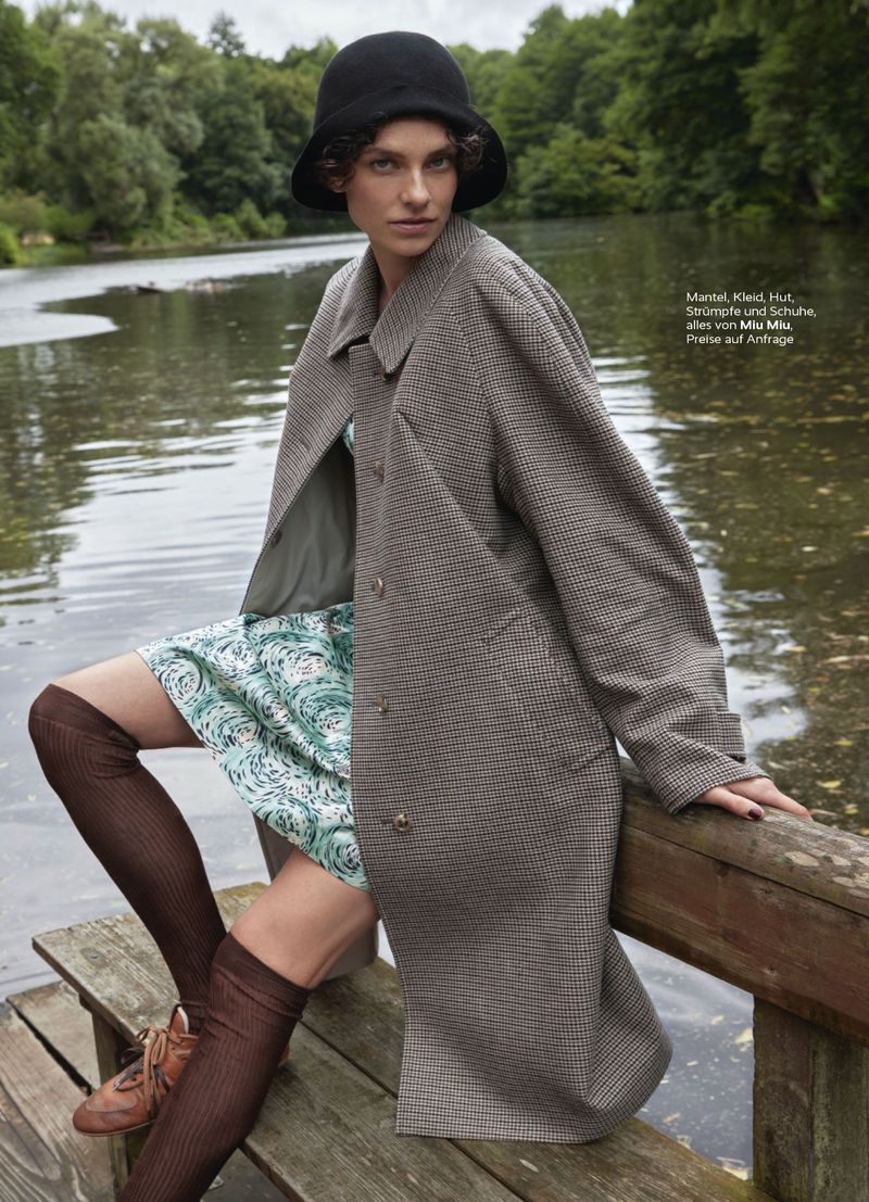 Woman in coat, hat, and knee-high socks sitting on a wooden rail near a lake surrounded by green trees in an outdoor fashion shot.