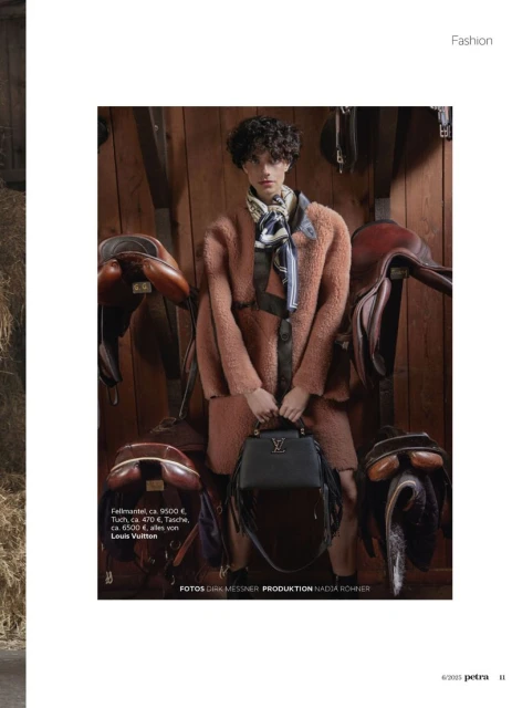Fashion shot of a woman in a fur coat holding a handbag, posed in front of saddles in a rustic, wood-paneled stable setting.