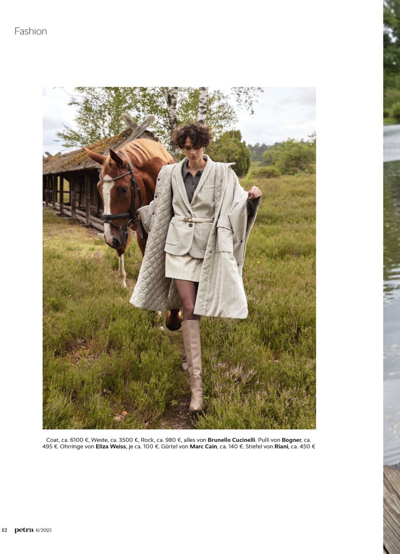 Model walking through an outdoor grassy field alongside a horse, wearing a light-coloured coat, skirt, and tall boots.