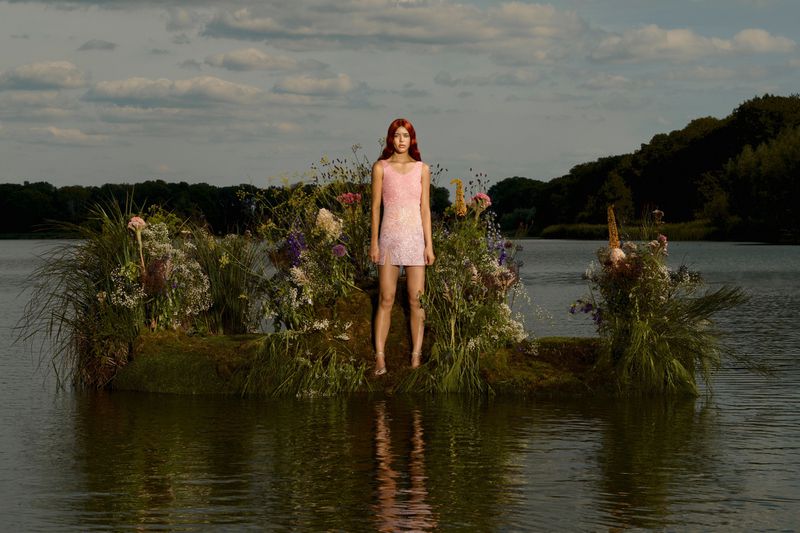 Woman in pink dress stands in a lake surrounded by wildflowers and grass. The sky is partly cloudy over a dark treeline.