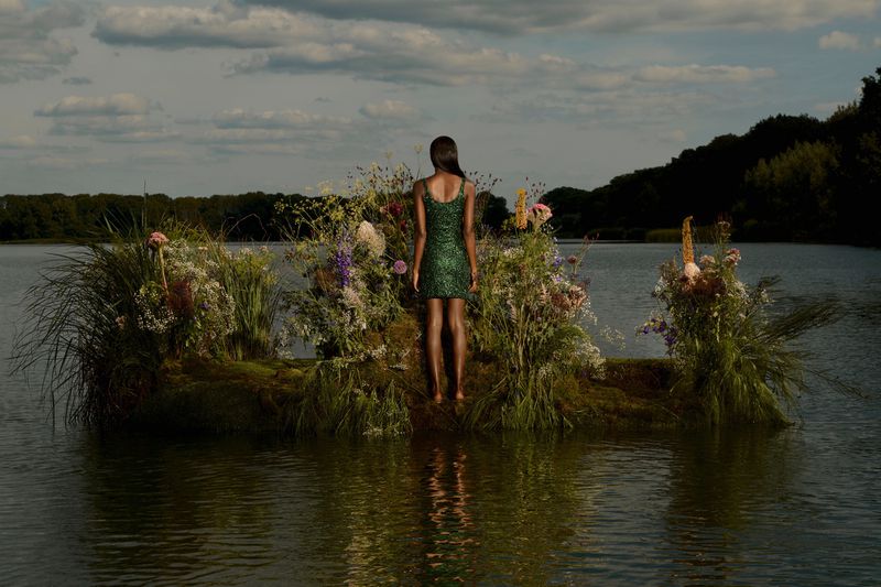 A woman in a short green dress stands on a grassy platform with flowers in a lake, facing away, under a cloudy sky.