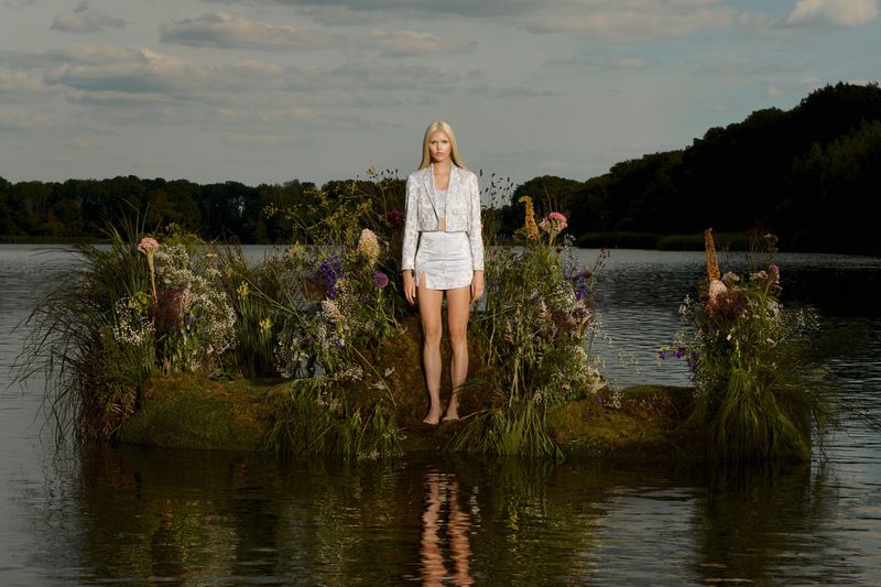 A woman stands barefoot on a grassy island in a lake wearing a matching white skirt and jacket in an outdoor fashion shot.