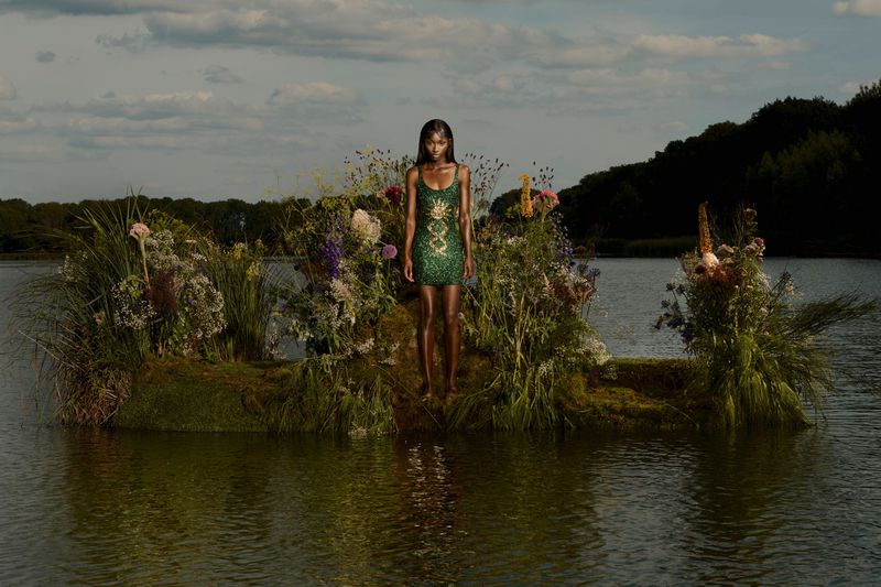 A woman in a short green dress stands on a mossy patch in a lake surrounded by flowers, under a cloudy blue sky, a nature scene.