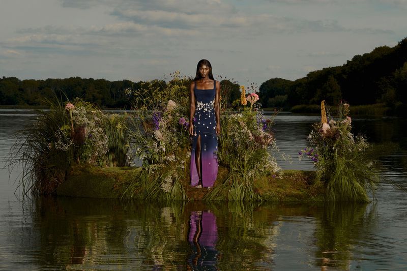 Woman in elegant gown stands on a grassy platform surrounded by water with a tree-lined horizon. Floral design fashion photography.
