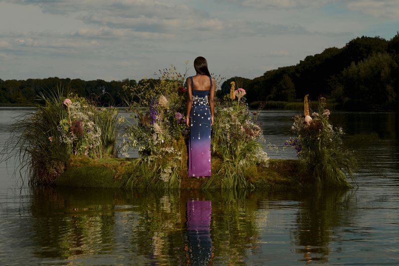 Elegant woman in formal gown stands on small grassy island surrounded by water and adorned with wildflowers, trees in the background.