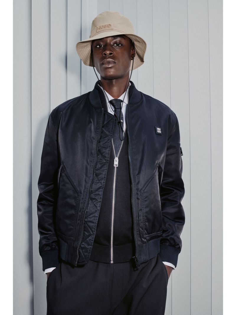 Dark-skinned male model in studio wearing a navy bomber jacket, bucket hat, tie, and black pants, stands against a blue-gray backdrop.