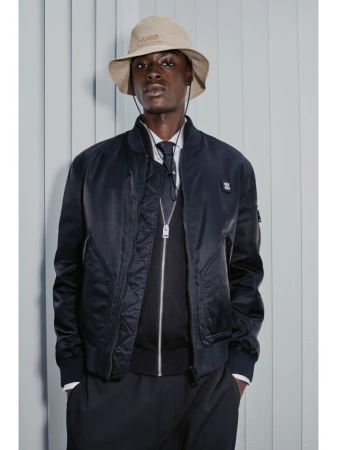 Dark-skinned male model in studio wearing a navy bomber jacket, bucket hat, tie, and black pants, stands against a blue-gray backdrop.