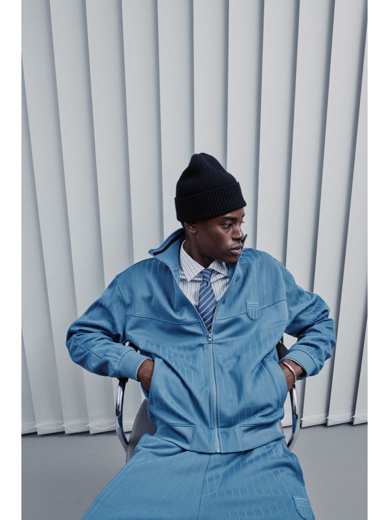 Portrait of African American man in black beanie, blue striped jacket and pants, sitting on chair against a light grey background.