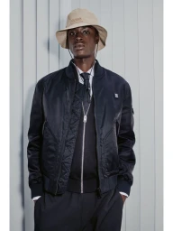 Babacar N'doye wearing a black bomber jacket and beige bucket hat against a vertically panelled studio backdrop for HUGO.