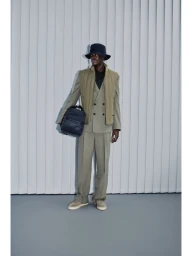 Model wearing an olive green suit, quilted vest, and bucket hat holding a dark backpack against a grey vertical-slat studio wall.