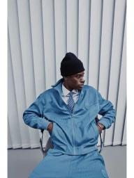 Model Babacar N'doye seated wearing a light blue tracksuit jacket, striped tie, and black beanie against vertical blinds.