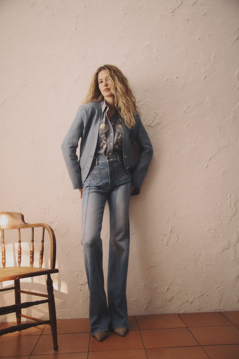 A stylish adult woman wearing denim jeans and a blazer poses in front of a textured wall, accessorized with a wooden chair.