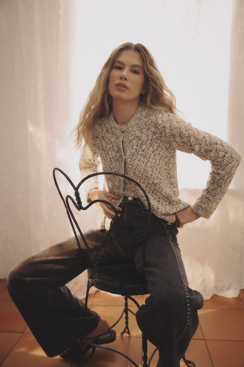 A blonde woman in a knitted jacket and dark jeans sits on a metal chair in a soft light setting, looking directly at the camera.