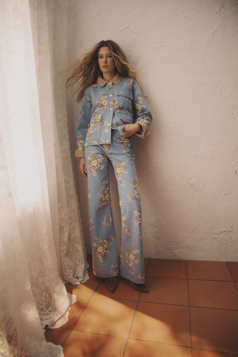 A woman stands indoors wearing a matching floral denim jacket and flared jeans. The light is soft and the background is simple.