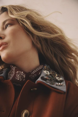 Close-up of a blonde woman in a fashionable rust-colored coat, adorned with beaded collar, showcasing beauty and high-end fashion details.