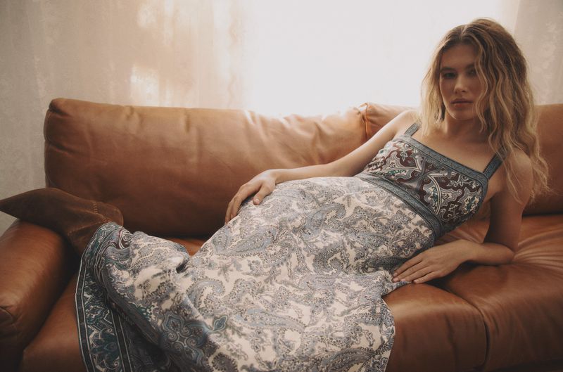 A woman with long blonde hair rests on a tan leather couch wearing a light-colored printed dress, relaxing indoors with soft, natural lighting.