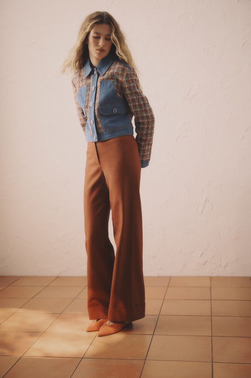 Fashion shot of a young adult woman posing in a jacket and wide-leg trousers against a peach-toned wall and terracotta tile floor.