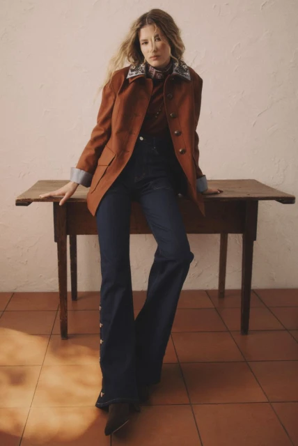 Woman in a rustic brown jacket, dark denim jeans, and brown boots poses next to a small wooden table in a warm indoor setting.