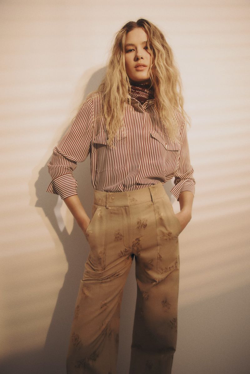 A long-haired blonde woman poses indoors wearing a striped shirt and patterned beige slacks. She is looking at the camera.