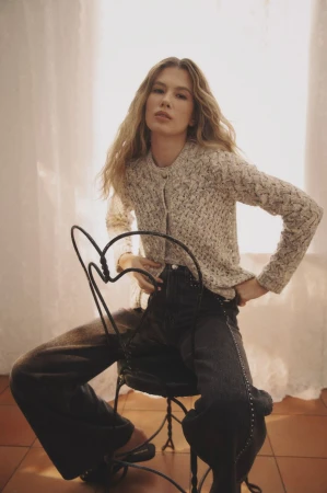 Model seated backwards on a black wrought-iron chair wearing a textured knit cardigan and dark wide-leg jeans.