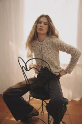 Model seated backwards on a black wrought-iron chair wearing a textured knit cardigan and dark wide-leg jeans.