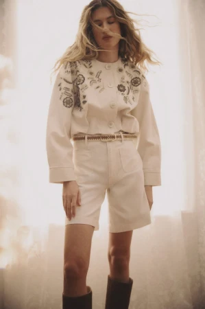 Model wearing embroidered cream jacket and matching shorts standing against a soft-lit window for Veronica Beard