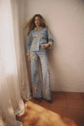 Model Meg Dmitruk in a light blue floral jacket and matching wide-leg trousers standing beside a lace curtain on terracotta tile.