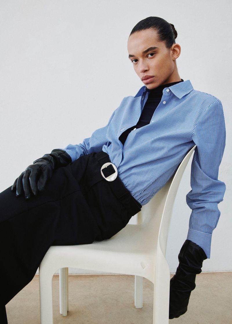 Person seated wearing a blue-striped shirt, black pants with a circular buckle, and long black gloves in a neutral indoor setting.